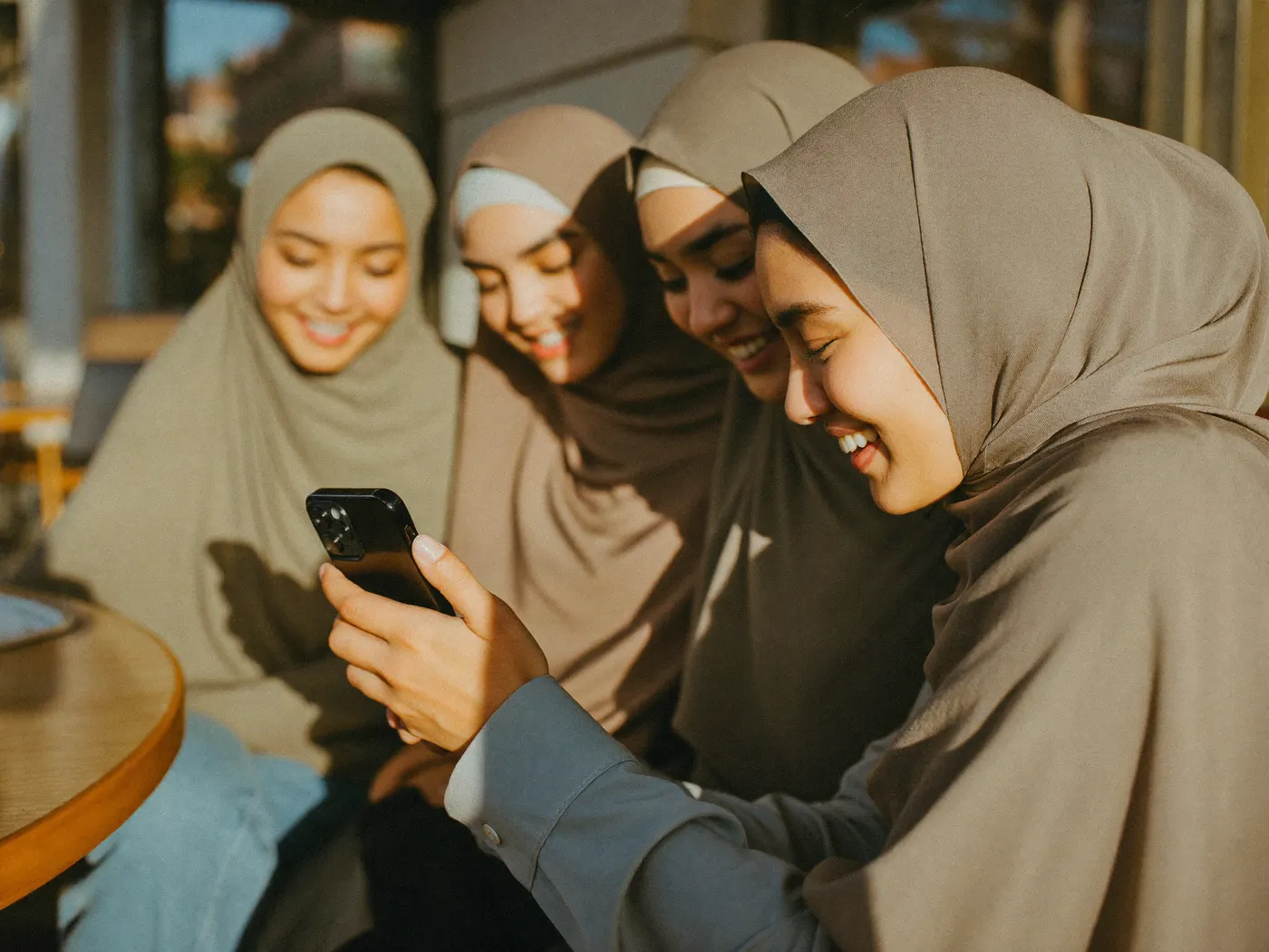 Four young women in hijabs sitting close together on a sunlit bench, all smiling at a phone one of them is holding.